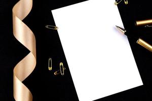 Gold pen, ribbon, paper clips and stationery on a black background with a white sheet of paper with copy space photo