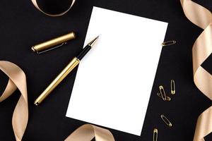 Gold pen, ribbon, paper clips and stationery on a black background with a white sheet of paper with copy space photo