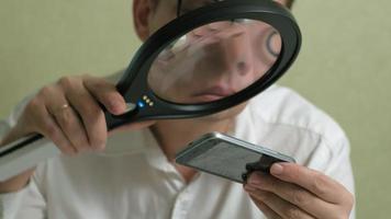 un hombre con gafas examina un teléfono con una pantalla táctil rota a través de una lupa video