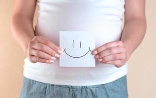 A close-up view of the belly of a pregnant woman holding a piece of paper with a smiley face photo