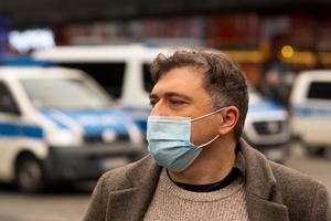 Retrato al aire libre de un hombre que protege su rostro con una máscara médica o quirúrgica mirando a un lado en el fondo de los coches de policía enfocados foto