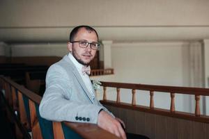 Foto de boda de las emociones de un novio barbudo con gafas en una chaqueta gris en el edificio de la iglesia