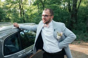 The groom with a beard in a gray jacket and glasses stands leaning on the car