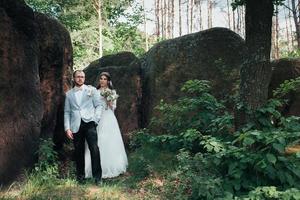 La novia y el novio en el día de la boda abrazándose de pie cerca de una roca o una piedra grande foto