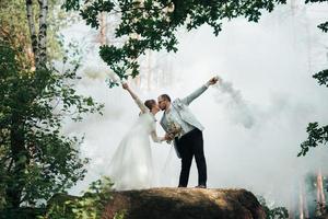 The bride and groom stand on a cliff and hold smoke bombs in their hands in white photo