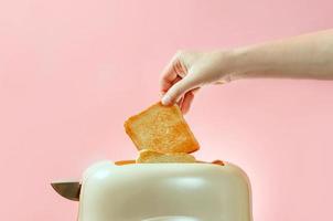 Una mano femenina saca un trozo de pan tostado de una tostadora sobre un fondo de color rosa con un espacio de copia foto
