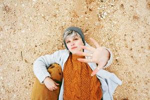 An adventurer young caucasian woman lying on grit ground beside a backpack wearing wool sweater and cap with her hand open and the orange as principal color photo