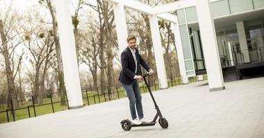 Hombre de negocios joven en ropa casual montando un scooter eléctrico por un edificio de oficinas en una reunión de negocios foto
