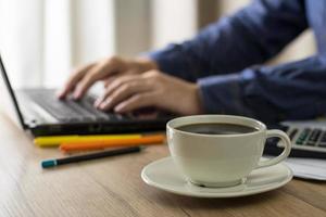 Taza de café con leche en el fondo de un hombre que trabaja con un portátil en casa y el empresario bebe café mientras trabaja foto