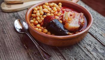 garbanzos, salchicha y tocino en una olla de cocción lenta con una cuchara de madera y un tenedor y una pequeña cuchara de metal foto