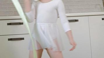 Une petite fille heureuse dans un maillot de bain de gymnastique blanc entraîne des danses avec un ruban pour des sauts de gymnastique rythmique et des exercices professionnels video