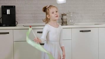 A happy little girl in a white gymnastic swimsuit trains dances with a ribbon for rhythmic gymnastics jumps and performing professional exercises