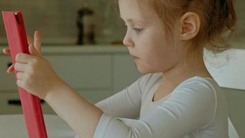 Jolie petite fille en vêtements blancs assis dans la cuisine à la table et jouer à l'aide d'une tablette, surfer sur le web, gros plan portrait video