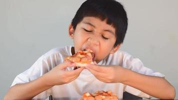 menino bonito asiático com camisa branca sentado feliz comendo pizza