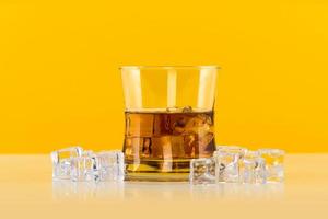 Glass of whiskey with ice cubes on yellow background photo