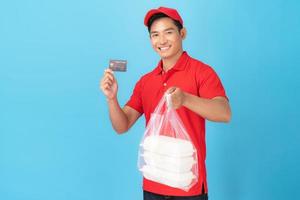 Smiling delivery man employee in red cap blank shirt uniform  standing with credit card giving food order