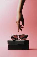 Hand reaching for a pair of sunglasses over a pastel pink background
