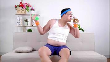 Funny fat Asian guy in sportswear lifting dumbbell weight training with enjoy drinking beer for refresh energy Happy overweight man workout and sitting on couch at living room while quarantine