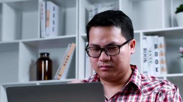 Portrait Asian glasses man looking to screen monitor with stressed face at home office in quarantine period Male using and typing on computer notebook with book shelf background video