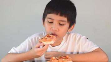 menino bonito asiático com camisa branca sentado feliz comendo pizza
