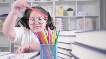 menina asiática de óculos está fazendo o dever de casa para a escola no deck branco, concentrando-se em seus desenhos e pinturas a lápis de cor em uma aconchegante sala de estar em sua casa depois de terminar as aulas