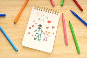 Cuaderno del concepto del día del padre con el dibujo de un padre con una niña y rotuladores sobre una mesa de madera foto