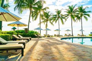 Hermosa sombrilla de lujo y una silla alrededor de la piscina al aire libre en el hotel y resort con palmera de coco en el cielo azul foto