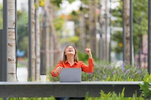 Hermosa mujer joven con cara de sorpresa gritando feliz usando la computadora portátil en un parque de la ciudad foto