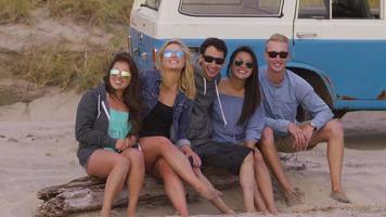 Retrato de amigos sentados en registro en la playa con vehículo en segundo plano. video