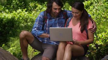 Pareja sentada sobre las rocas durante la caminata y usando una computadora portátil video