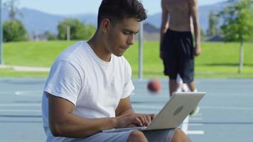 Joueur de basket-ball adolescent à l'aide d'un ordinateur portable à la cour extérieure video