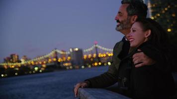 Pareja en la ciudad de Nueva York de pie en el muelle por la noche con el horizonte de la ciudad en segundo plano. video