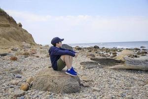 Boy sitting on a rock on the beach looking at the ocean photo