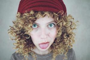 Closeup retrato de una bella y divertida mujer joven con ojos azules y cabello rubio rizado sacando la lengua con un gorro de lana rojo foto