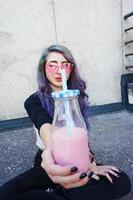 Feliz hermosa adolescente con gafas de sol rosa aclama y disfruta de una bebida rosa sentada en suelo urbano foto