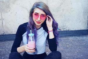 Feliz hermosa adolescente con gafas de sol rosa bebe y disfruta de una bebida rosa sentada en suelo urbano foto