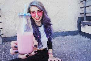 Feliz hermosa adolescente con gafas de sol rosa aclama y disfruta de una bebida rosa sentada en suelo urbano foto