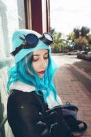 Retrato de una mujer joven punk o gótica con cabello de color azul y vistiendo gafas steampunk negras y gorro de lana azul en una calle urbana al aire libre foto