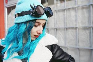 Retrato de una mujer joven punk o gótica con cabello de color azul y vistiendo gafas steampunk negras y gorro de lana azul en una calle urbana al aire libre foto