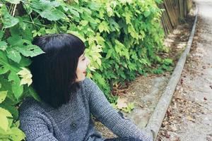 Beautiful caucasian brunette woman sitting on ground in a park wearing punk or gothic clothes and surrounded by green leafs photo