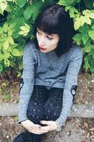 Beautiful caucasian brunette woman sitting on ground in a park wearing punk or gothic clothes and surrounded by green leafs photo