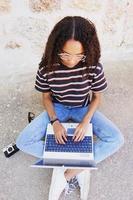A portrait of focused young black woman with curly hair wearing glasses, jeans and a striped t-shirt, next to technology as smartphones and cameras, sitting on the ground and working or making homework photo