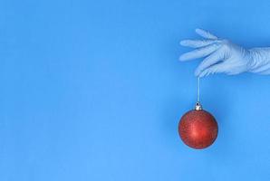 La mano en el guante médico sostiene la bola de Navidad roja sobre fondo azul con espacio de copia.