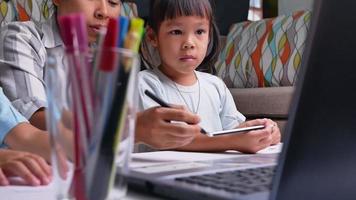 feliz madre e hijas aprendieron a dibujar a través de la computadora portátil en la sala de estar. madre ayudando a las niñas a hacer un dibujo en papel. concepto de escuela en casa. clases en línea de jardín de infantes. video