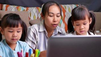 glückliche Mutter und Töchter lernten, durch den Laptop-Computer im Wohnzimmer zu zeichnen. Mutter hilft den Mädchen, ein Bild auf Papier zu zeichnen. Schulkonzept zu Hause. Online-Kurse im Kindergarten.
