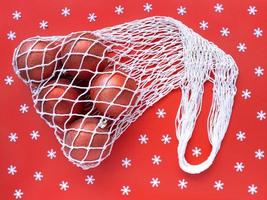 White string cotton eco bag with five christms tree balls on red background and scattered white snowflakes. Holiday simple flat lay. Ecology zero waste concept. Stock photo. photo