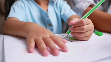 Nahaufnahme Hand des kleinen Mädchens lernte, durch den Laptop-Computer im Wohnzimmer zu zeichnen. Mutter hilft dem Mädchen, ein Bild auf Papier zu zeichnen. Schulkonzept zu Hause. Online-Bildung im Kindergarten.