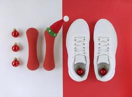 plano de vacaciones deportivas. bolas de Navidad, zapatillas y mancuernas con lindo sombrero sobre un fondo blanco y rojo. foto