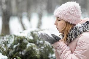 Mujer feliz en un día nevado de invierno en el parque, vestida con ropa de abrigo, sopla la nieve de sus guantes