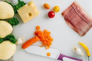 Sliced carrot with knife, potato, cheese, bacon, some onion and herbs on table. photo